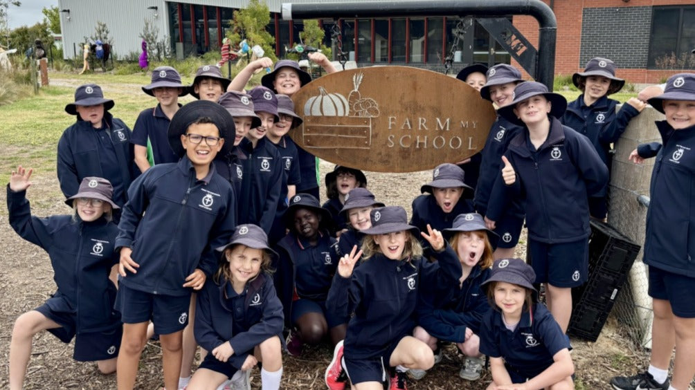 A group of smiling primary school students gathered around the Farm My School sign, celebrating real food and garden-based learning.